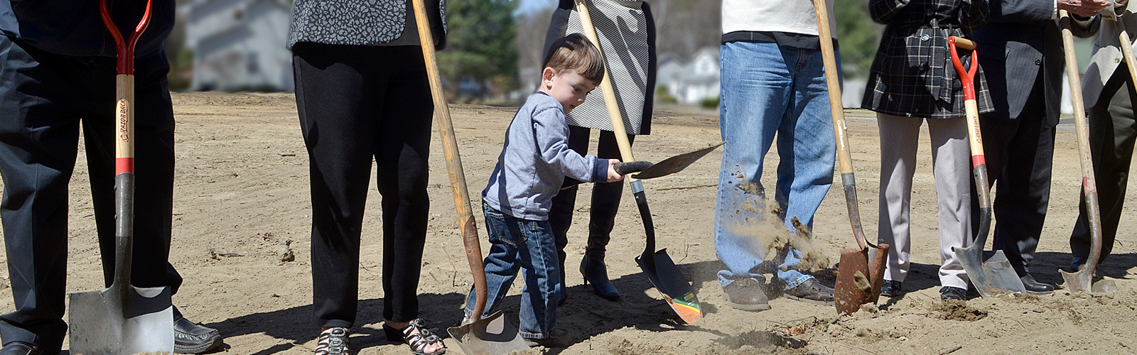 Three adults' legs and one child digging dirt with shovels at a ground breaking.