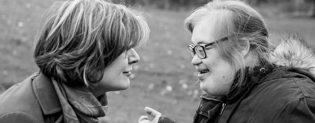 Woman with short hair smiling and looking at a young woman with Down syndrome, who is smiling and looking at the woman with short hair. Grass and trees in the background.