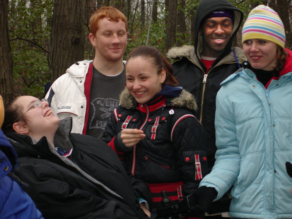 Five young adults with coats on smiling together. One young woman is laughing leaning back, looking at the other young adults. Trees in the background.